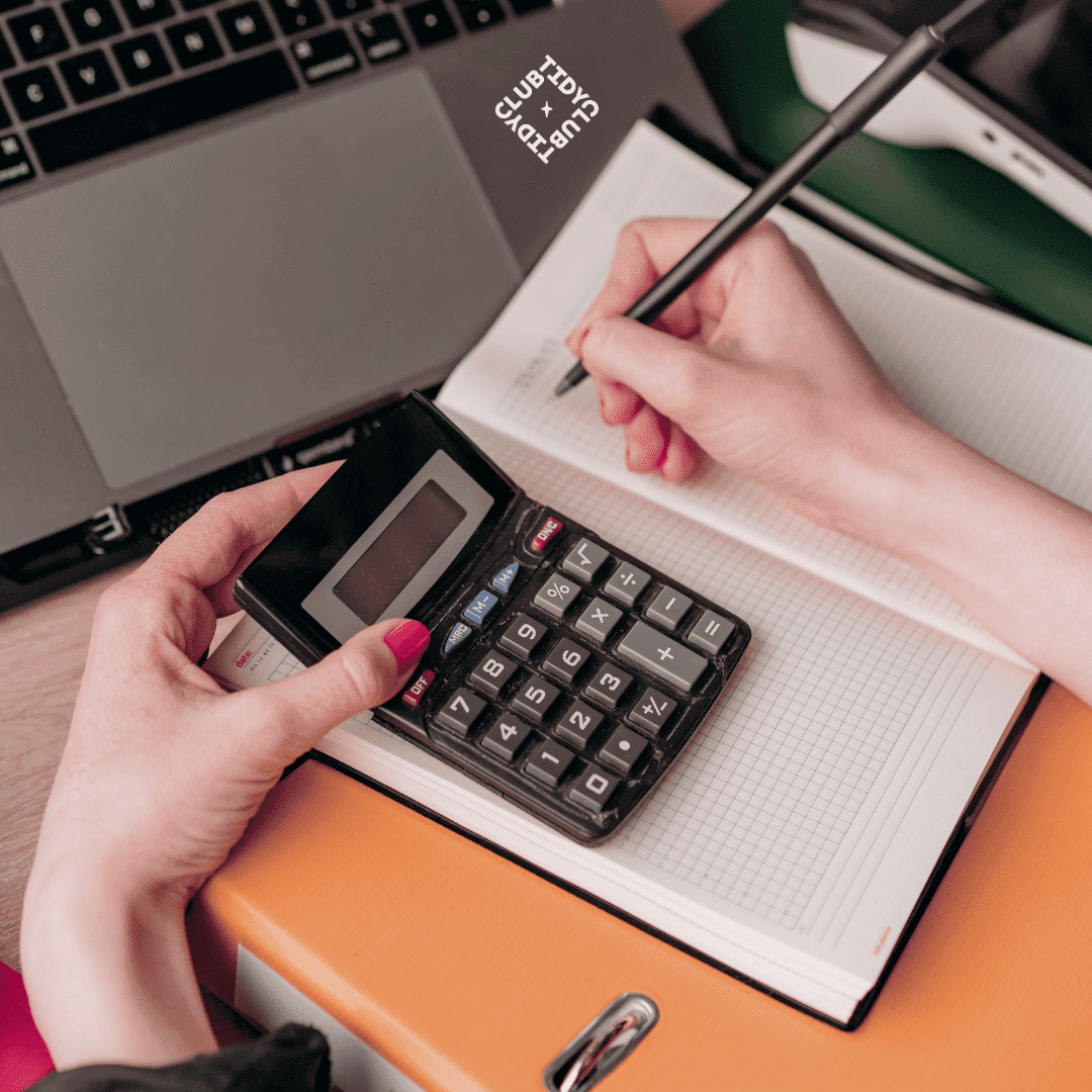 Photo d'une personne tenant une calculatrice et écrivant dans un cahier, avec un ordinateur portable en arrière-plan.