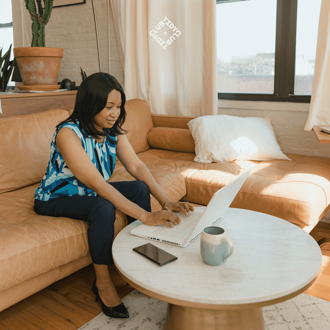 Femme assise sur un canapé, utilisant un ordinateur portable sur une table basse avec un téléphone et une tasse à côté.