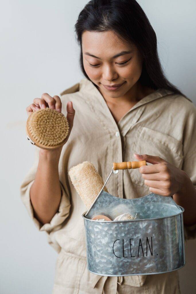 Femme tenant un seau en métal marqué 'CLEAN' contenant des brosses et des accessoires de nettoyage.